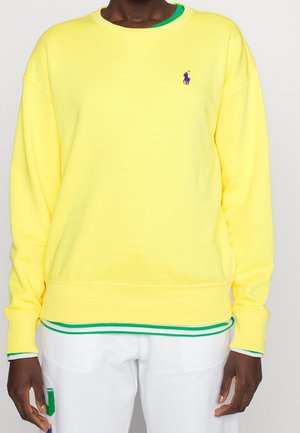 Osoba ubrana w żółtą bluzę z kapturem z małym fioletowym logo na piersi, pod którą ma koszulę z zielonym obszyciem oraz białe spodnie z zielonymi i niebieskimi naszywkami.