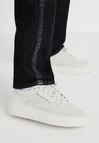 White suede sneakers with perforated detailing and a chunky sole, paired with dark denim jeans featuring a light vertical seam.