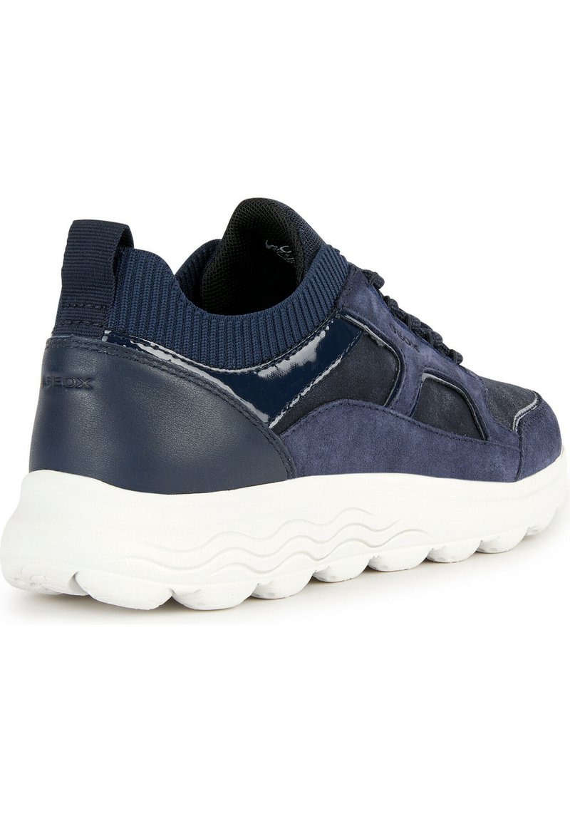 Zapatilla azul marino con una combinación de ante y charol, que cuenta con un cuello de punto texturizado y una suela de goma blanca ondulada.