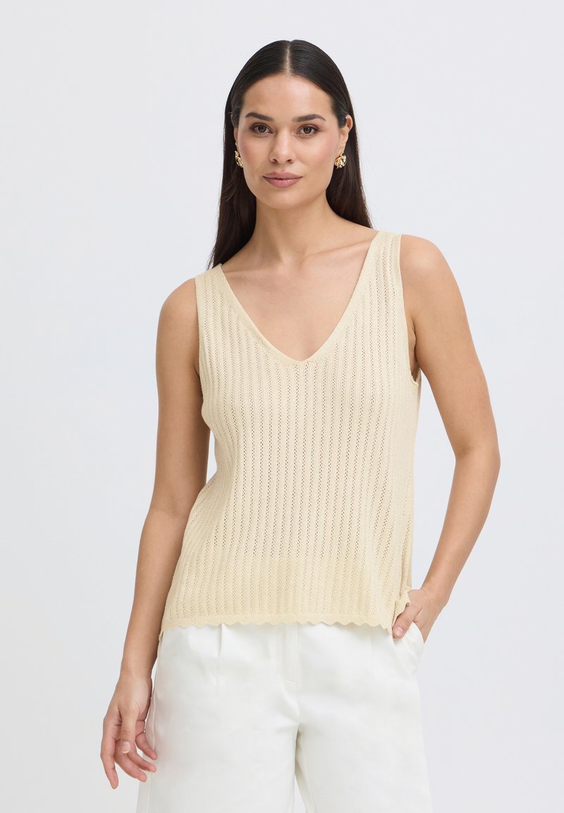 Femme aux cheveux raides et foncés portant des boucles d'oreilles en or, un haut sans manches en maille beige et un pantalon blanc, debout avec une main dans la poche.