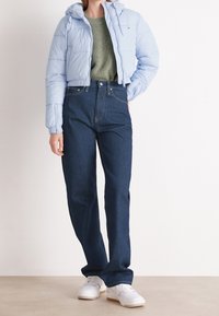 Osoba v světle modrém vypodloženém kabátu, zeleném pleteném svetru, tmavě modrých kalhotách s vysokým pasem a bílých teniskách stojí na béžové podlaze.