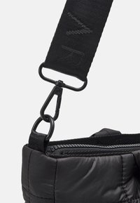 Czarna poduszka torba w rogu z suwakiem, przymocowana do szerokiego czarnego paska z widocznym szyciem i czarnym metalowym hakem.