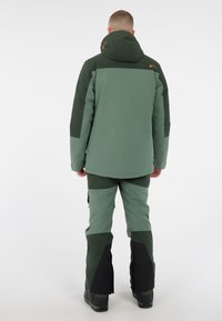 Grüne isolierte Jacke mit Kapuze, die ein zweifarbiges Design aufweist. Kombiniert mit passenden Hosen, die verstärkte Unterschenkel und schwarze Akzente zeigen.