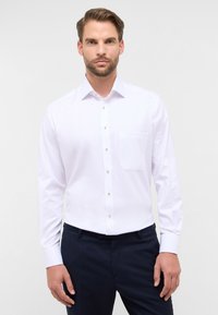 Mann in einem weißen langärmligen Hemd mit Knopfleiste und Brusttasche, dazu dunkle marineblaue Hose, steht vor einem einfarbigen hellen Hintergrund.