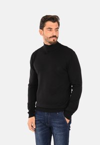 Maglione nero a collo alto realizzato in materiale morbido, con polsini e orlo a coste, abbinato a jeans di denim blu. Il modello si trova in piedi con le mani in tasca.