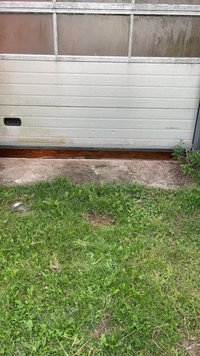 Κλειστή, φθαρμένη λευκή πόρτα γκαράζ με σκουριασμένη κάτω άκρη, επι PATCH τσιμέντου στην είσοδο και πράσινο γρασίδι με μερικά ζιζάνια εμπρός.