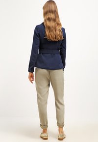 Námořnicky modré sako na míru s páskem, kombinované s lehce béžovými zúženými kalhotami a zlatými nazouvacími botami. Vyznačuje se hladkou texturou a elegantním designem.