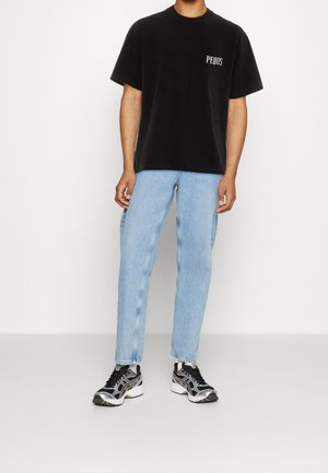 Uomo che indossa una maglietta nera con logo PEQUA, jeans azzurri chiari, calzini bianchi e scarpe da ginnastica nere, bianche e argento in piedi contro uno sfondo bianco.