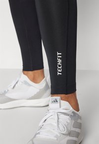 Leggings deportivos negros con un ajuste ceñido, que presentan un logo blanco de "TECHFIT" en el costado. Combinados con unas zapatillas deportivas blancas y grises.