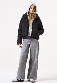 Schwarze Pufferjacke mit Kapuze, versehen mit einem gesteppten Design und Reißverschlusstaschen, kombiniert mit lockeren grauen Jeans und weißen Sneakers.