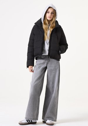 Doudoune noire avec capuche, adoptant un design matelassé et des poches zippées, associée à un jean en denim gris ample et des baskets blanches.