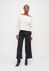 Schulterfreies weißes Sweatshirt mit geripptem Kragen, kombiniert mit schwarzen, gestreiften weiten Hosen und schwarzen, spitzen Ankle Boots.