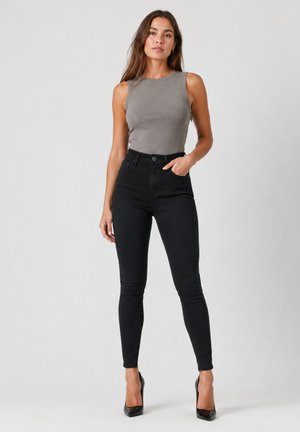 Donna con capelli castani lunghi che indossa un top grigio senza maniche, jeans skinny neri a vita alta e tacchi alti neri, in piedi davanti a uno sfondo semplice.