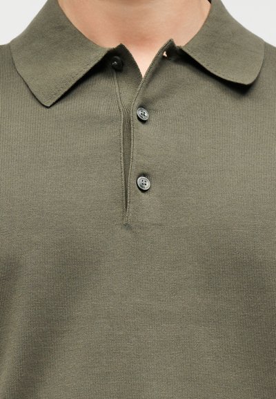 Κοντινό πλάνο πλεκτού πουκάμισου πόλο σε ελιά πράσινο με τρία κουμπιά, το πάνω κουμπί ανοιχτό, φορεμένο από ένα άτομο.