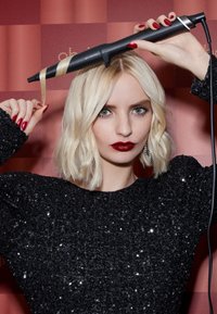 Fer à friser noir maintenu au-dessus des cheveux blonds mi-longs, avec un pull noir texturé brillant, des ongles rouges et un arrière-plan doucement éclairé.