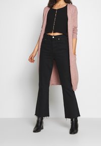 Zwarte flared jeans met rafelige zoom, gedragen met een geribbelde zwarte crop top en een lichtroze lange cardigan met zakken, met zichtbare knoopdetails.