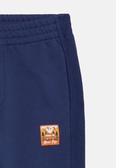 Námornícky modré tepláky s textúrovaným povrchom, ktoré majú oranžovú vyšitú nášivku znázorňujúcu hory a karavan s textom "Cesta za dobrodružstvom".