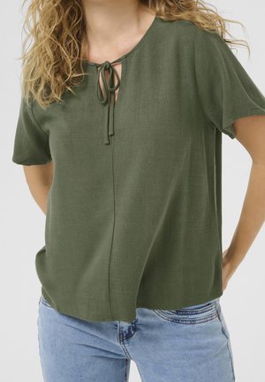 Kobieta nosząca oliwkową bluzkę z krótkim rękawem i wiązaniem z przodu przy dekolcie, w połączeniu z jasnoniebieskimi dżinsami, na neutralnym tle.