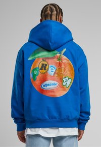 Sudadera de algodón azul con un gran gráfico naranja en la parte posterior que presenta varios logotipos coloridos y un detalle de hoja verde. Puños y dobladillo con canalé.