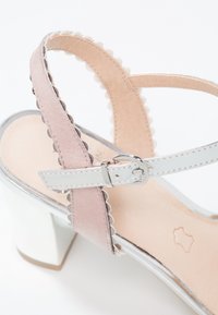 Sandale à bout ouvert avec bride de cheville en cuir blanc et boucle argentée, bride à bord festonné rose, et talon épais blanc.