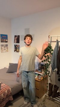 Mladý človek v bežnom oblečení zdraví v spálni s posteľou, násteným umením, nočným stolíkom, rastlinami a stojanom na oblečenie s čižmami a bundami.