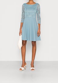 Robe bleu clair avec des manches en dentelle trois-quarts et un corsage ajusté. Jupe évasée avec une texture lisse. Sandales à talons blanches et un petit sac.