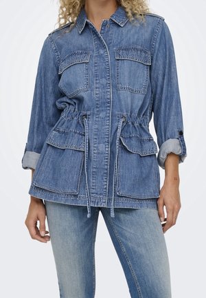Kurtka dżinsowa z czterema klapkowymi kieszeniami, ściągaczem w talii i podwiniętymi rękawami, noszona przez osobę w jasnoniebieskich dżinsach.