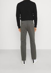 Mężczyzna w ciemnoszarych spodniach, czarnej długiej koszulce i białych trampkach stoi twarzą do gładkiej białej ściany.