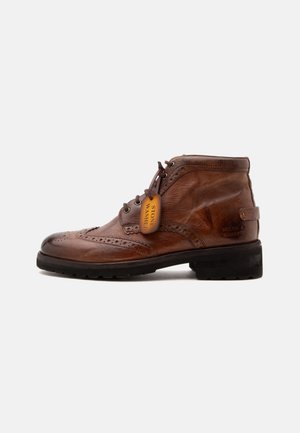 Bota de tobillo brogue de piel marrón con cordones, suela negra y una etiqueta "Stone Washed" adjunta a los cordones sobre un fondo blanco.