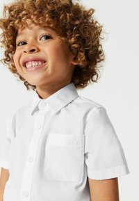 Chemise blanche à manches courtes avec un col pointu, une fermeture à boutons, et une poche sur la poitrine gauche avec un logo en relief. Texture de tissu lisse.