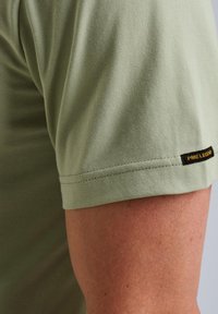 Olivovozelené tričko s jemnou textúrou, viditeľným šitím a čiernou etiketou so žltým textom na ľavom rukáve.