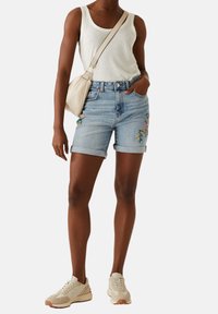 Top in tessuto leggero grigio chiaro, pantaloni shorts di jeans blu con ricami floreali e sneakers beige. Una borsa a tracolla in un tono chiaro completa l'outfit.