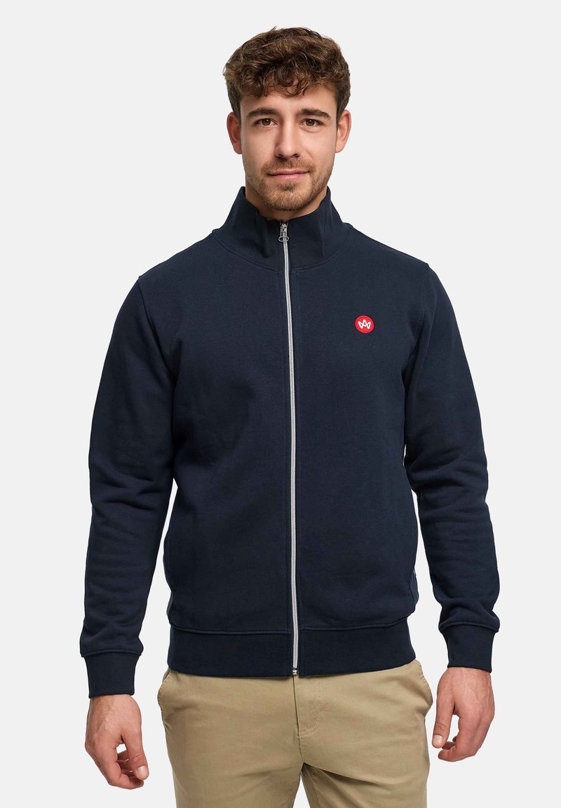 Sweat-shirt zippé bleu marine en tissu doux, doté d'un col haut, de poignets côtelés et d'un logo rouge sur la poitrine. Texture lisse et coupe ajustée.