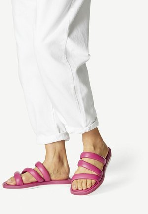 Personne portant un pantalon blanc retroussé et des sandales slides roses avec trois brides rembourrées sur des pieds nus, sur fond blanc.