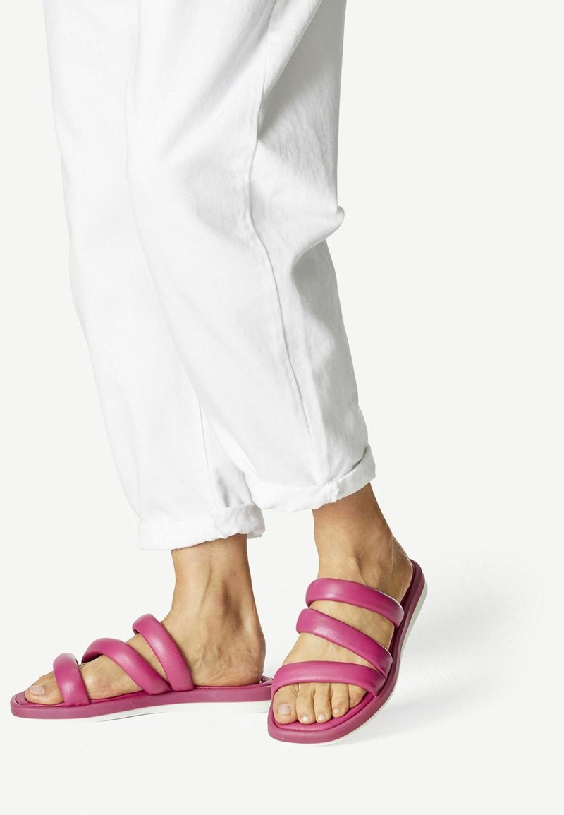 Personne portant un pantalon blanc retroussé et des sandales slides roses avec trois brides rembourrées sur des pieds nus, sur fond blanc.