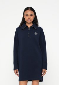 Robe sweat-shirt bleu marine oversize avec col zippé, manches longues, et petit logo brodé sur la poitrine. Texture de tissu doux.