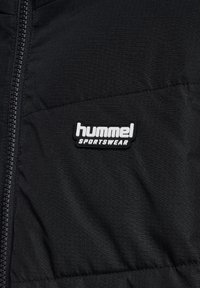 Sort sportsjakke med tekstureret overflade og lynlåslukning. Har et hvidt logo patch med teksten "hummel SPORTSWEAR" på brystet.