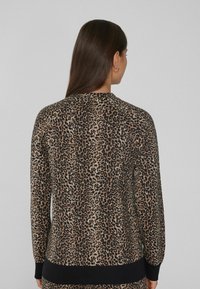 Žena s dlouhými tmavými vlasy má na sobě halenku s leopardím potiskem a černými manžetami, otočená zády k jednobarevnému světle šedému pozadí.