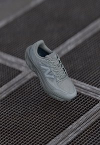Svijetlosivi New Balance tenisice za trčanje s pamučnom gornjom i debelim đonom, smještene iznad metalne rešetke s međusobno presijecanjem linija.