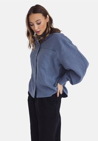 Niebieska lniana koszula z guzikami, z jedną kieszenią na piersi, luźny krój i obszerne rękawy. Noszona z czarnymi spodniami, prezentując swobodny look.