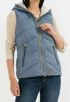 Personne portant une veste sans manches matelassée bleue avec capuche, une chemise beige à manches longues et un jean bleu, avec une main en train de fermer la veste à glissière.