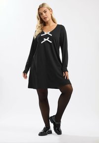 Schwarzes langärmliges Kleid mit V-Ausschnitt, zwei weißen Schleifenakzenten auf der Brust und einer lässigen Passform. Getragen mit schwarzen Strumpfhosen und Schuhen.