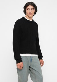 Mladý človek s krátkymi kučeravými vlasmi, oblečený v čiernom textúrovanom svetri cez bielu košeľu a voľných svetlomodrých džínsoch, ktorý sa pozerá na stranu.