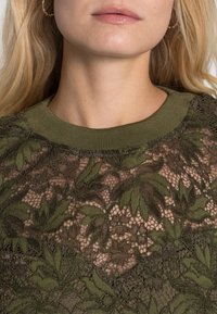 Olivgrön spetstopp med intrikata blommönster, med en ribbad halsringning och genomskinlig textur som framhäver lager av bladdesigner.