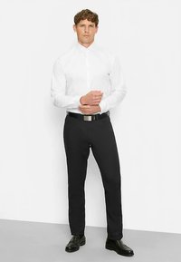 Weißes Langarmhemd, schwarze Hose, schwarzer Gürtel mit silberner Schnalle und schwarze elegante Schuhe, Modell steht mit verschränkten Händen, neutraler Hintergrund.