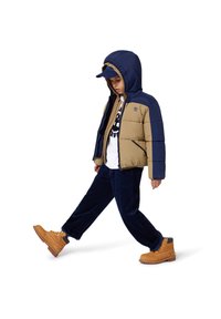 Giacca imbottita in blu navy e khaki, con cappuccio e zip. Abbinata a pantaloni in velluto a coste scuri e stivali beige con lacci.