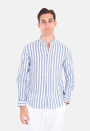 Uomo con camicia bianca a maniche lunghe con righe verticali blu e pantaloni bianchi, in piedi davanti a uno sfondo chiaro e uniforme.