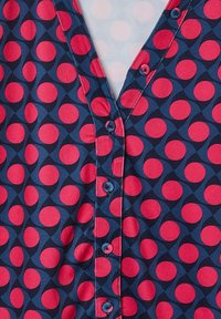 Red and navy patterned blouse featuring large circles, a V-neckline, and a button-down front. Soft fabric with a smooth texture.