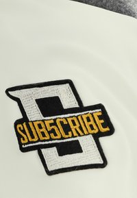 Biely, hladký materiál obsahuje čierne a žlté vyšívané logo s nápisom "PREDPLATTE" a geometrické dizajnové prvky.