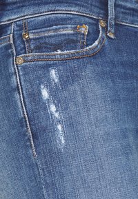 Jeans de mezclilla azul con un diseño ligeramente desgastado, con desgarros y marcas de abrasión visibles, acentuados por costuras amarillas contrastantes.